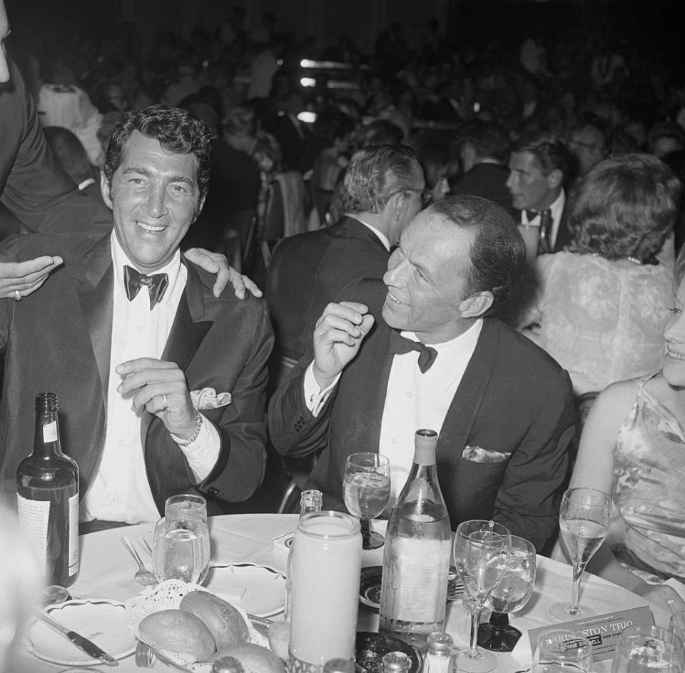 Dean Martin and Frank Sinatra in formal tuxedos sit laughing at a crowded event table with drinks and food