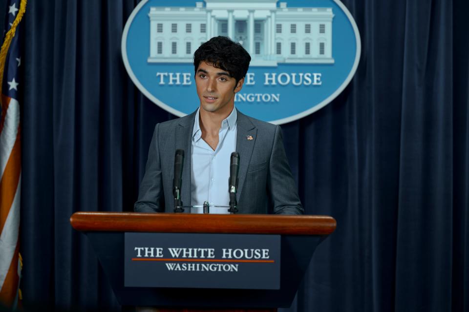 alex in red white and royal blue, dressed in a grey suit and shirt with the top button undone, speaking at a white house podium in the press room