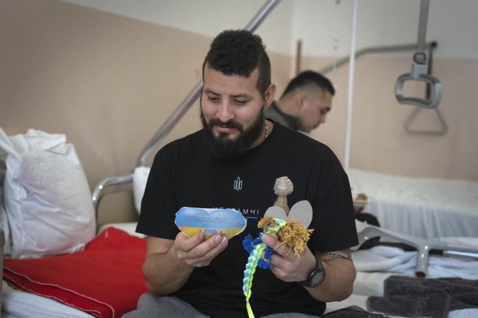 A wounded professional soldier from Medellín, Colombia who goes by the call sign of Checho, 32, smiles as he holds gifts from Ukrainian children in a hospital in Ukraine Wednesday, Dec. 20, 2023. Checho joined the Ukrainian armed forces to help fight Russia. After two years of war, Ukraine is looking for ways to replenish its depleted ranks. The Colombian professional soldiers are a welcome addition. (AP Photo/Efrem Lukatsky)