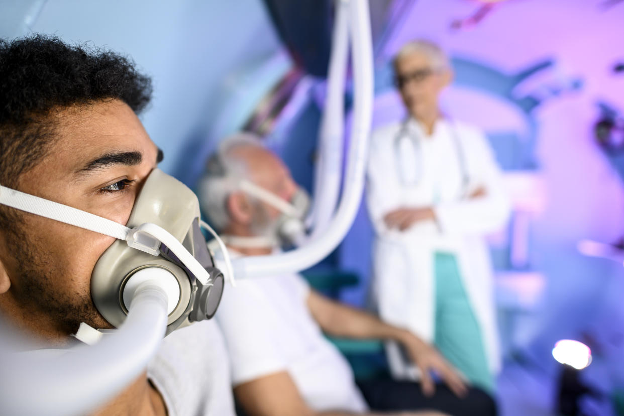 A multi-ethnic young patient holding a breathing mask, with another patient in the background.