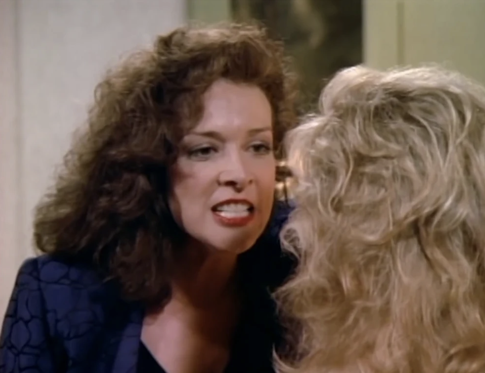 A woman with curly hair is intensely shouting at another woman with blonde hair in a scene from a film or TV show