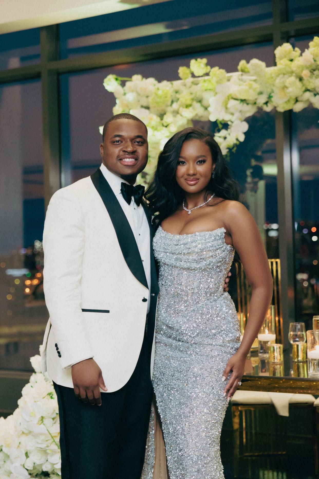 A bride and groom smile in wedding attire in front of a floral arch.