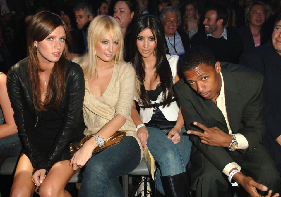 Three young women and a man holding a peace sign sit front row at a fashion show