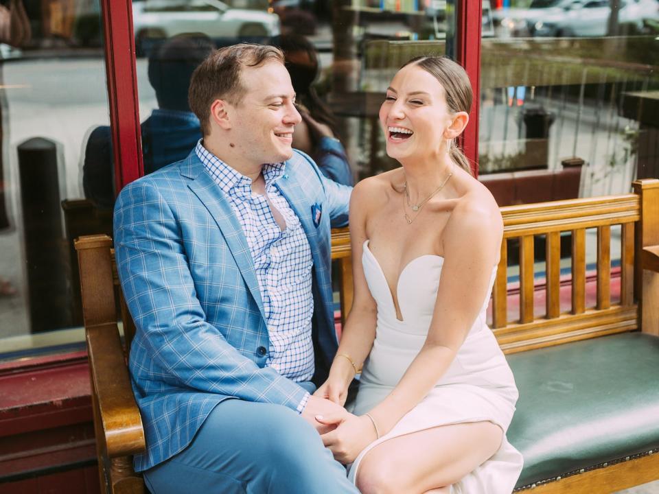 A bride and groom laugh on a bench.