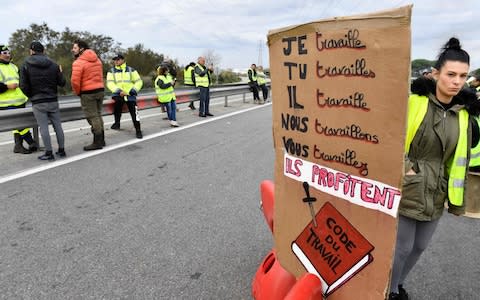 Yellow Vests (Gilets jaunes) standing near a sign reading "We work, they benefit" as they block the road during a demonstration against the rising of the fuel and oil prices on November 19, 2018 near the oil depot of Fos-sur-mer, southern France - Credit: GERARD JULIEN/ AFP