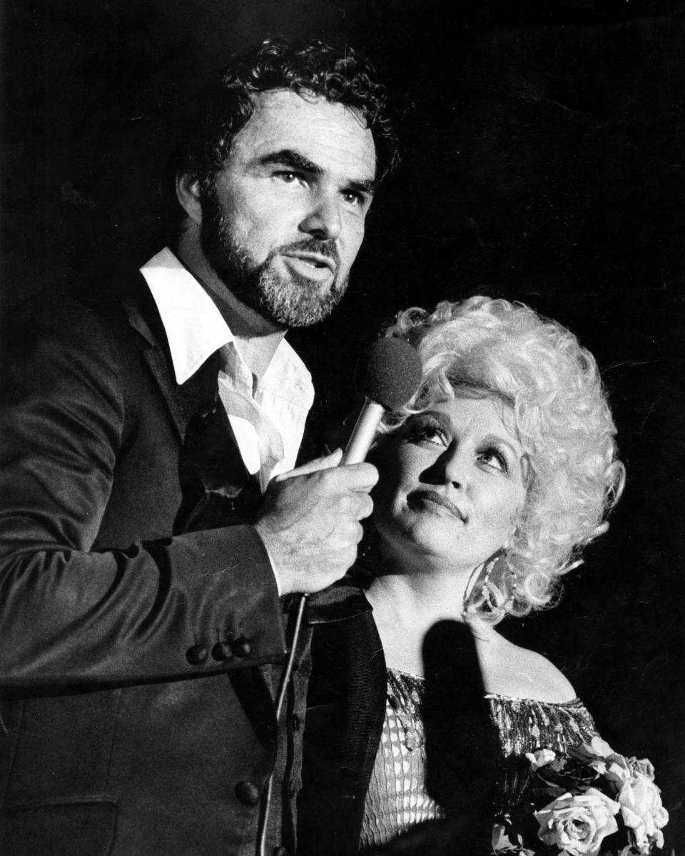 Black and white photo of Dolly Parton looking up to Burt Reynolds who is speaking into a microphone.