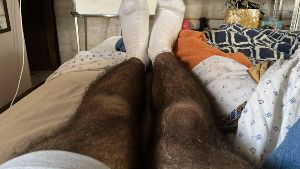 View from someone's perspective showing their legs in white socks, lying on a bed with multiple blankets and an unorganized room in the background