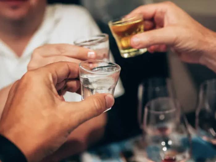 Three unidentified people clink shot glasses in a toast. The focus is on the hands and glasses; background details are blurred