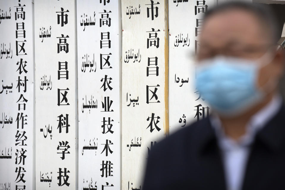 A government official stands near a sign board outside a location that was identified in early 2020 as a re-education facility by an Australian think tank, which the Chinese government asserts is currently home to a veterans' affairs bureau and other offices, in Turpan in western China's Xinjiang Uyghur Autonomous Region during a government organized trip for foreign journalists, Thursday, April 22, 2021. A spokesperson for the Xinjiang region called accusations of genocide "totally groundless" as the British parliament approved a motion Thursday that said China's policies amounted to genocide and crimes against humanity. (AP Photo/Mark Schiefelbein)