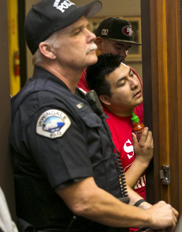 An Irwindale police officer, left, guards a door to a city council meeting, as a worker peeks holding a bottle of Sriracha hot sauce waits in the overflowed lobby in Irwindale, Calif., Wednesday, April 23, 2014. The city of Irwindale is suing the maker of Sriracha hot sauce and last week the Los Angeles suburb tentatively voted to declare the bottling plant a public nuisance. Irwindale City Attorney Fred Galante, not seen, says relocating seems extreme and the city only wants to see the smell issue addressed. (AP Photo/Damian Dovarganes)