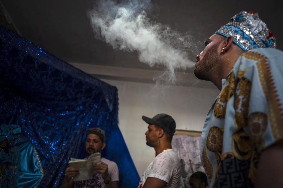 Mandy Arrazcaeta blows smoke standing in front of a blue altar during a Santería ceremony at his home in Havana, Cuba, Sunday, Nov. 13, 2022. Arrazcaeta, a white Cuban and member of the LGBTQ+ community, found refuge in the religion when he was 12. Once an Evangelical Christian, he said he felt rejected by members of that religion for being gay. (AP Photo/Ramon Espinosa)