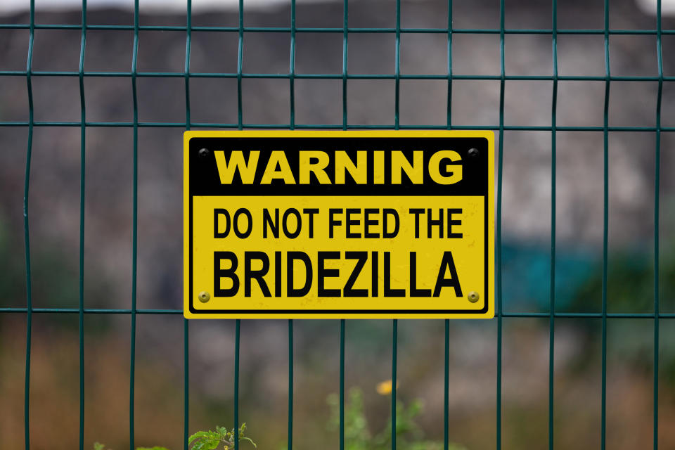 A yellow and black sign posted on a wire fence that reads "Warning Do Not Feed The Bridezilla."