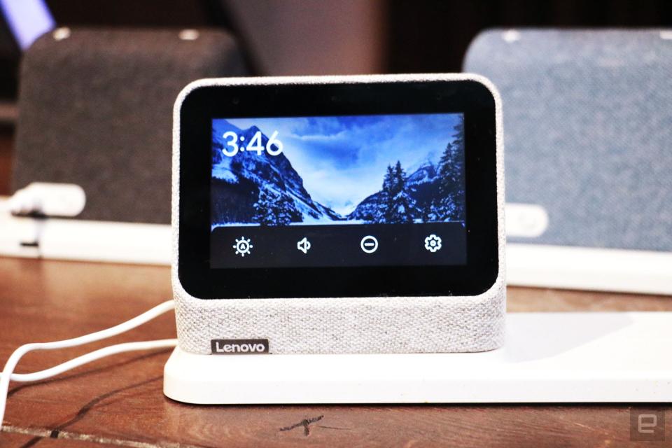 <p>A gray Lenovo Smart Clock 2 on a wireless charging dock with its screen facing the camera. A row of options for volume, brightness, do not disturb and settings take up the bottom of the display.  In the background are a black and a blue Smart Clock 2 on the left and right respectively.</p>
