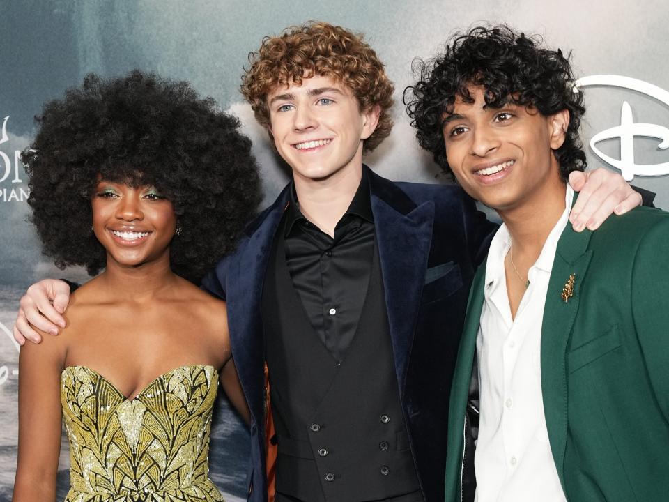 leah sava jeffries, walker scobell, and aryan simhadri together at the percy jackson premiere. jeffires is waring a yellow and blue patterned dress, scobell is in a navy jacket and vest, and simhadri is in an emerald green suit