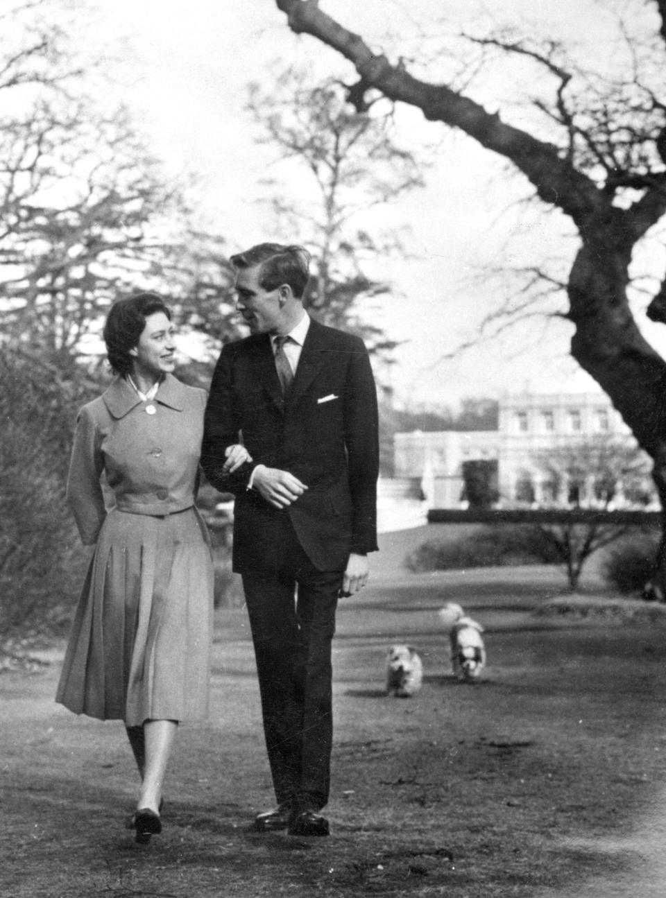 The newly-engaged Princess Margaret and photographer Antony Armstrong-Jones, strolling in the grounds of Royal Lodge, Windsor, where they are spending the weekend with the Queen Mother. This is one of the first pictures made of the couple after the announcement of their engagement. Princess Margaret wears a lilac dress with loose-fitting jacket, two strings of pearls and a gold watch. Her fiance's suit is a dark grey pin-stripe.