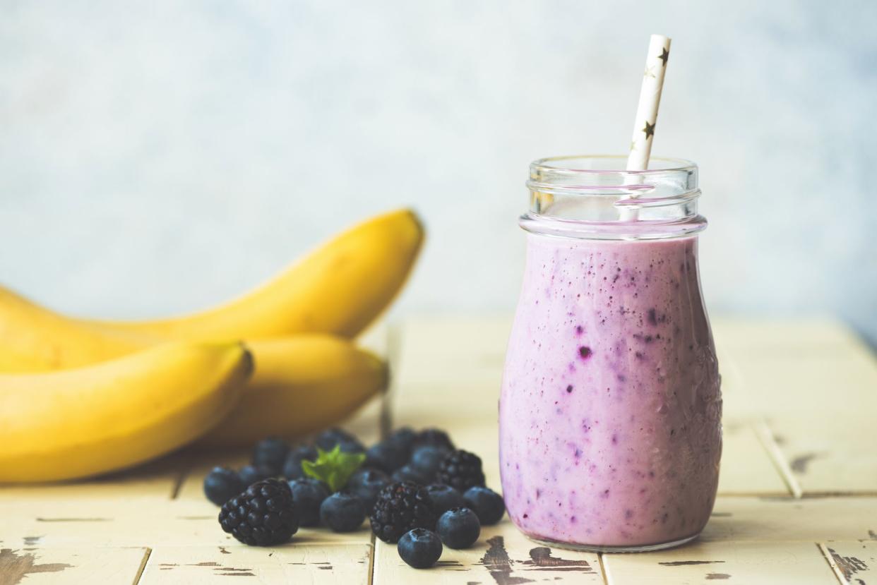 A blueberry and banana smoothie in a glass jar with a white straw with stars, next to blueberries and bananas, on a beige painted table, with a blurred background of a light blue wall