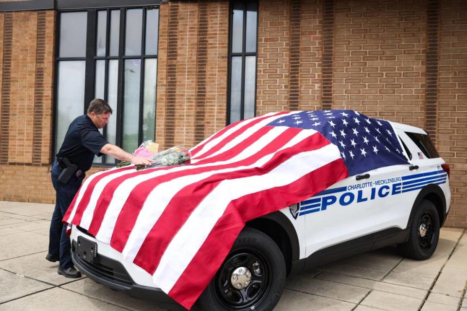 A CMPD officer adjusts flower bouquets on a police vehicle that draped in an American flag outside the police station on North Tryon on Tuesday, April 30, 2024. Flowers have been placed on the hood in memory of CMPD officer Joshua Eyer, who was killed in yesterday’s deadly shootout in East Charlotte.