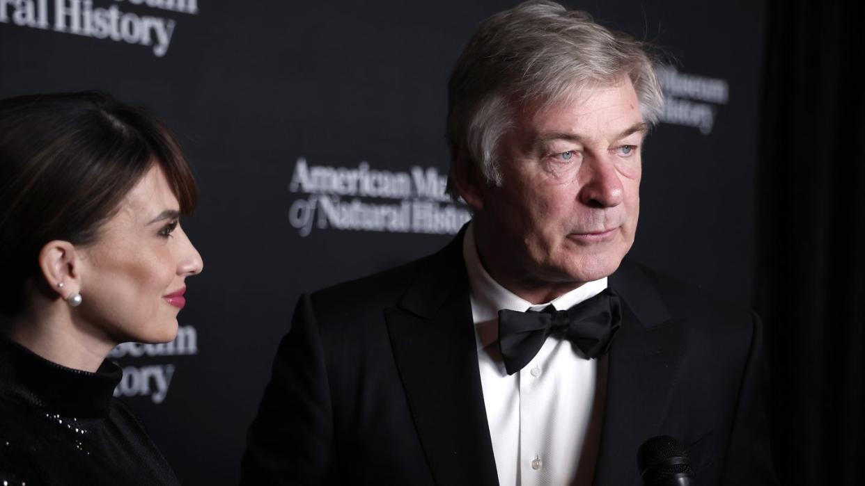 alec baldwin looks right while standing in front of a black photo background, he wears a tuxedo with a while collard shirt