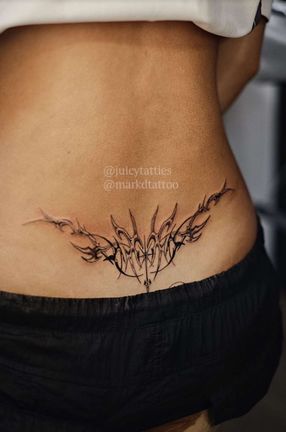 Lower back tattoo featuring an artistic abstract design with the text "@juicytatties @markdtattoo" written above the tattoo