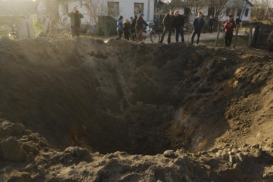 People look at a crater of an explosion in a village of Horodnya, Chernihiv region, Ukraine, Thursday, April 14, 2022. The fluid nature of the conflict, which has seen fighting shift away from areas around the capital and heavily toward Ukraine's east, has made the task of reaching hungry Ukrainians especially difficult. (AP Photo/George Ivanchenko)