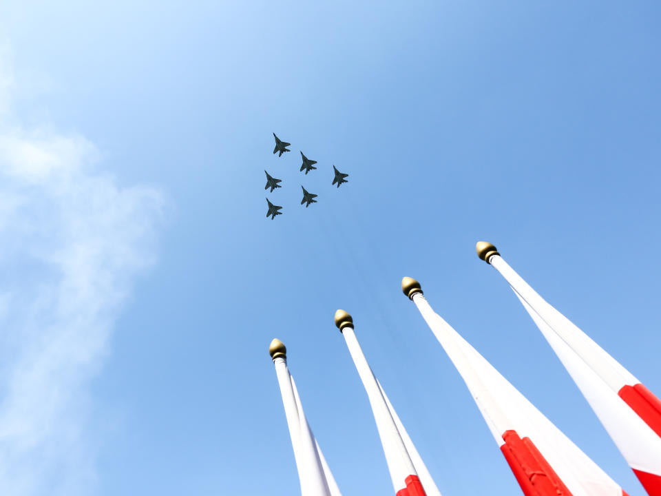 Polish Air Force MiG-29 aircraft fly in an arrow formation above four flagpoles with the Polish flag. 