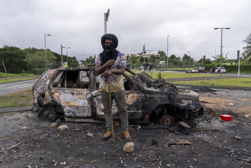 A man in New Caledonia stands in front of a burned out car on May 14, 2024.<br> - Photo: NICOLAS JOB/SIPA (AP)