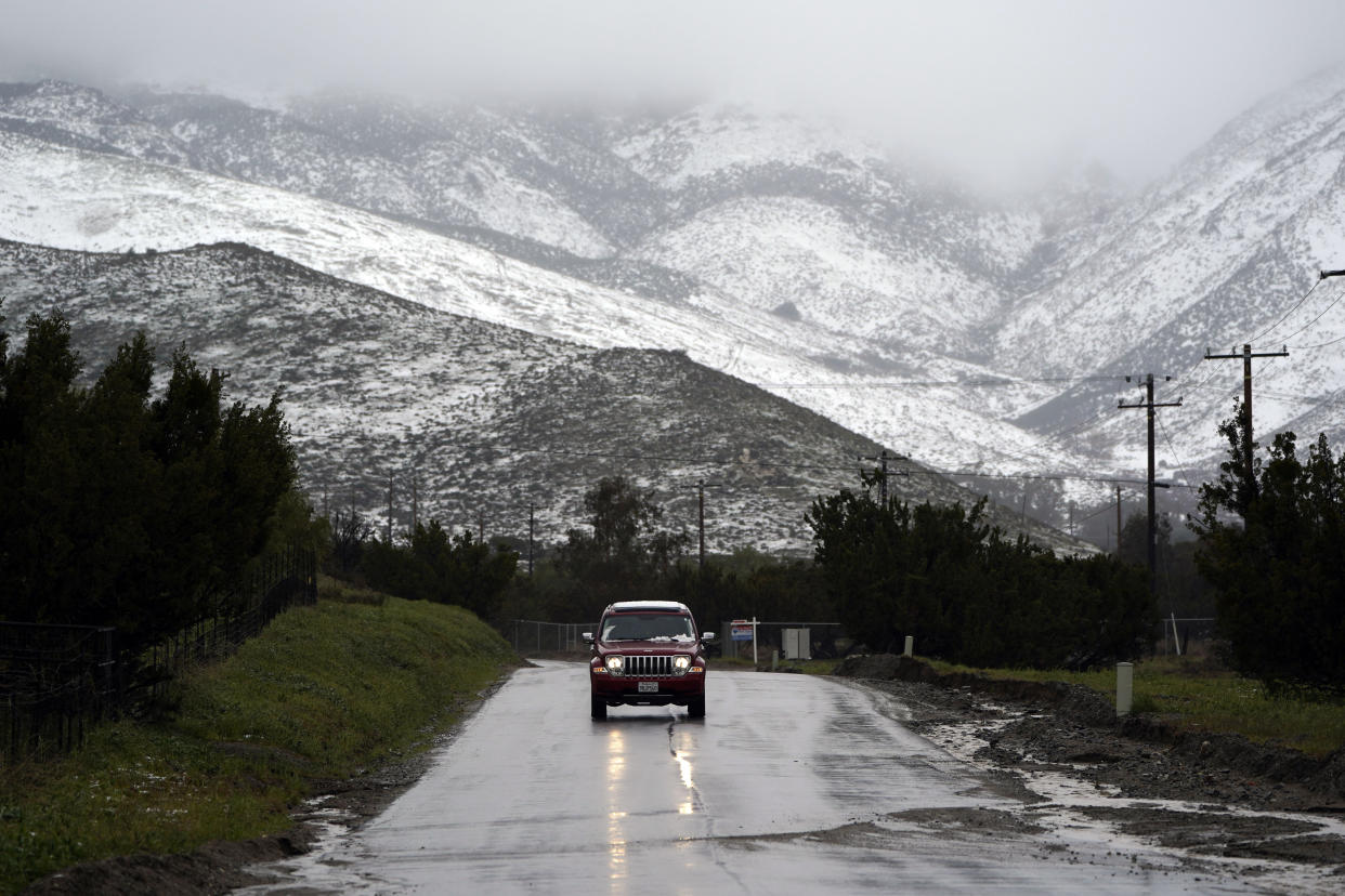 A motorist drives on a wet road under a snow-covered hillside Friday, Feb. 24, 2023, in Agua Dulce, Calif. California and other parts of the West faced heavy snow and rain Friday from the latest winter storm to pound the U.S. (AP Photo/Marcio Jose Sanchez)
