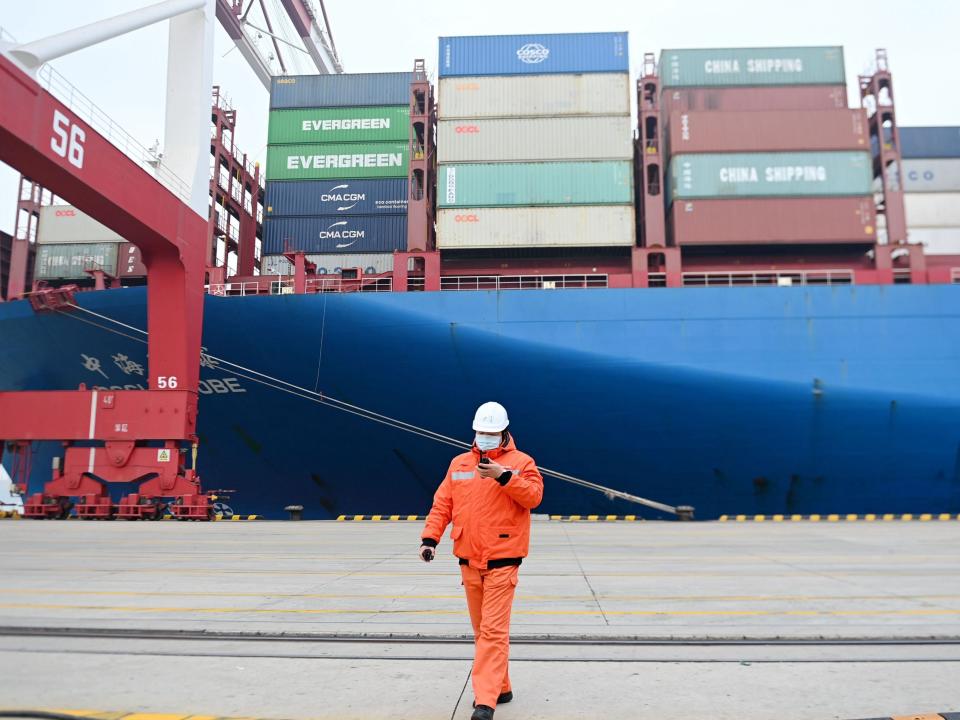 A masked man in orange in front of the side of a container ship in China.