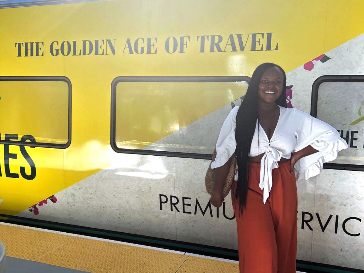 Christina Jane in a white tie top and reddish orange pants smiling in front of a Brightline train exterior