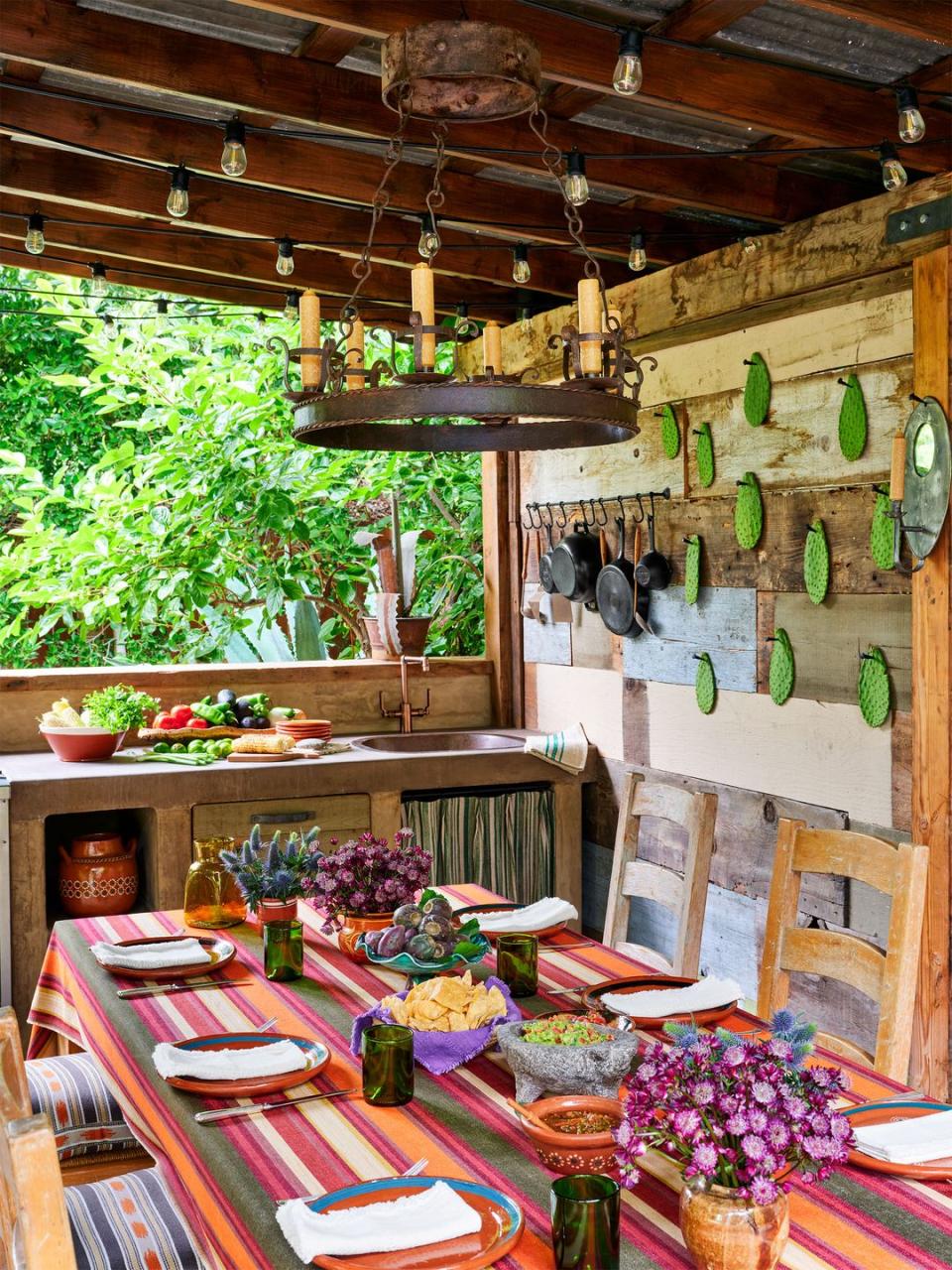 outdoor kitchen with cast iron skillets hanging on rustic wood wall, sink and counter with produce on top and cabinets below, set dining table with bright striped cloth and flowers, wood chairs with patterned seat cushions