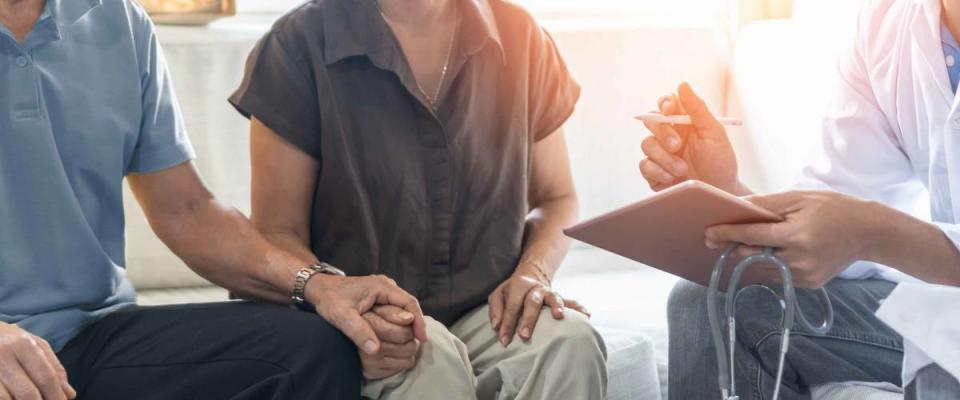 Elderly couple, sitting with doctor, holding hands while doctor writes on clipboard