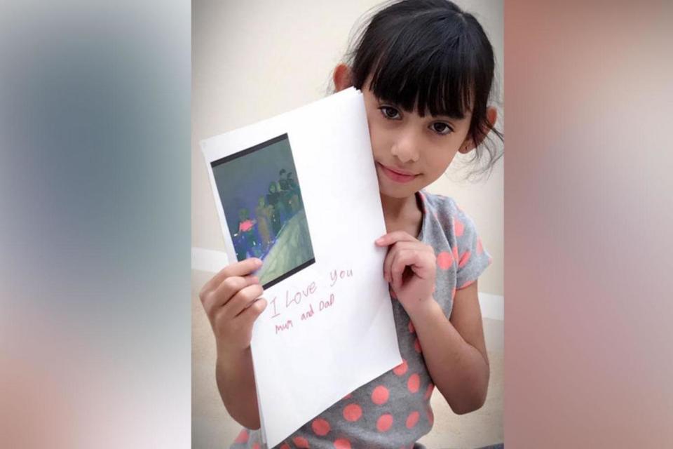 Sophia pictured smiling and holding a homemade card for her parents bearing the message: