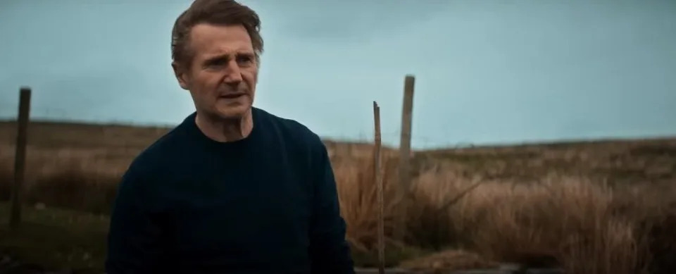 liam neeson in the land of saints and sinners movie, his character stands in front of a field and looks concerned