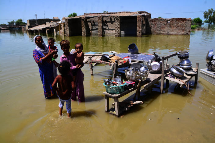 A woman and four children stand in water in a flooded area, next to wooden tables piled with gathered-up belongings.