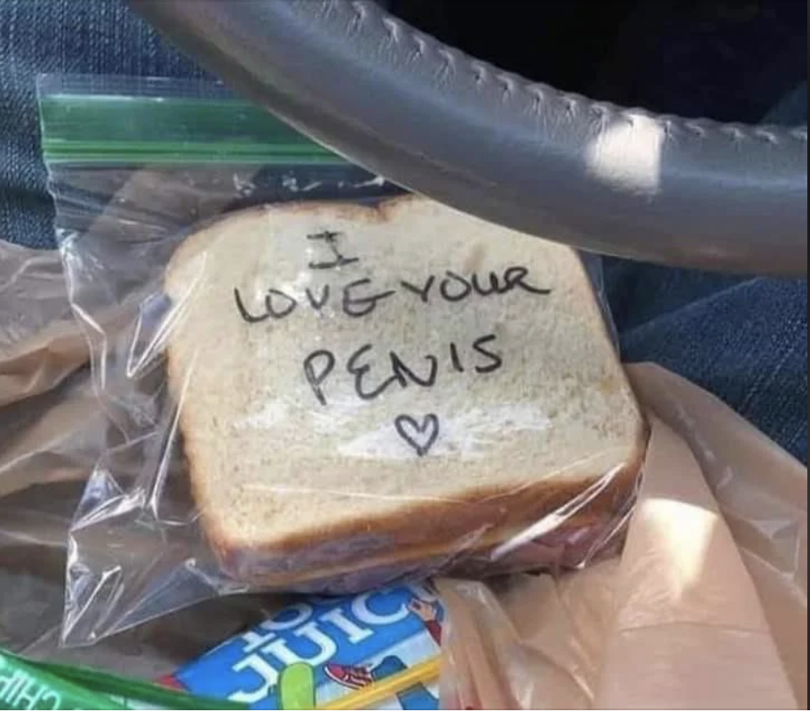 A sandwich in a plastic bag with text written on the bread: "I love your penis" and a heart symbol. The sandwich is on a car seat. Nearby, there's a juice box