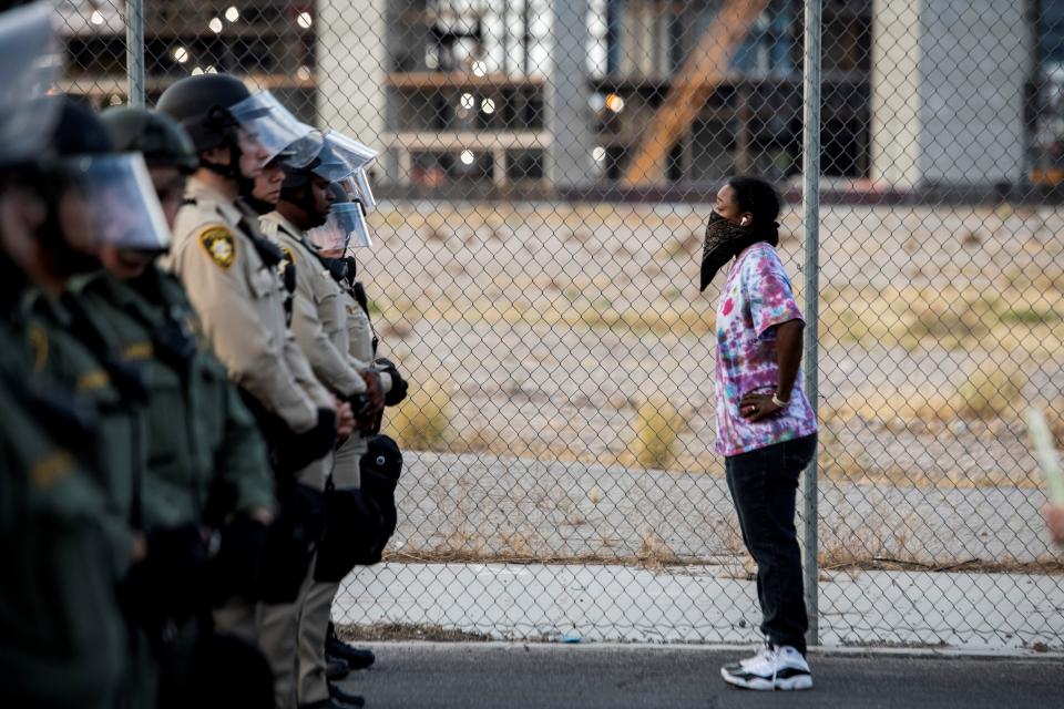 A Black woman wearing a face mask stands in front of Police officers with her hands on her hips