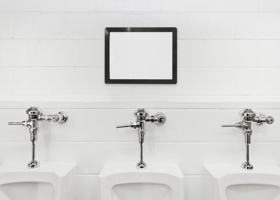 Three urinals are aligned under a blank, framed screen on a tiled wall