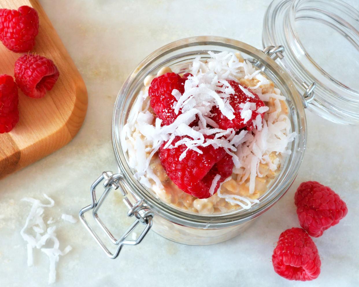 Top view of coconut milk oatmeal in a glass jar, surrounded by raspberries and coconut flakes on a white table