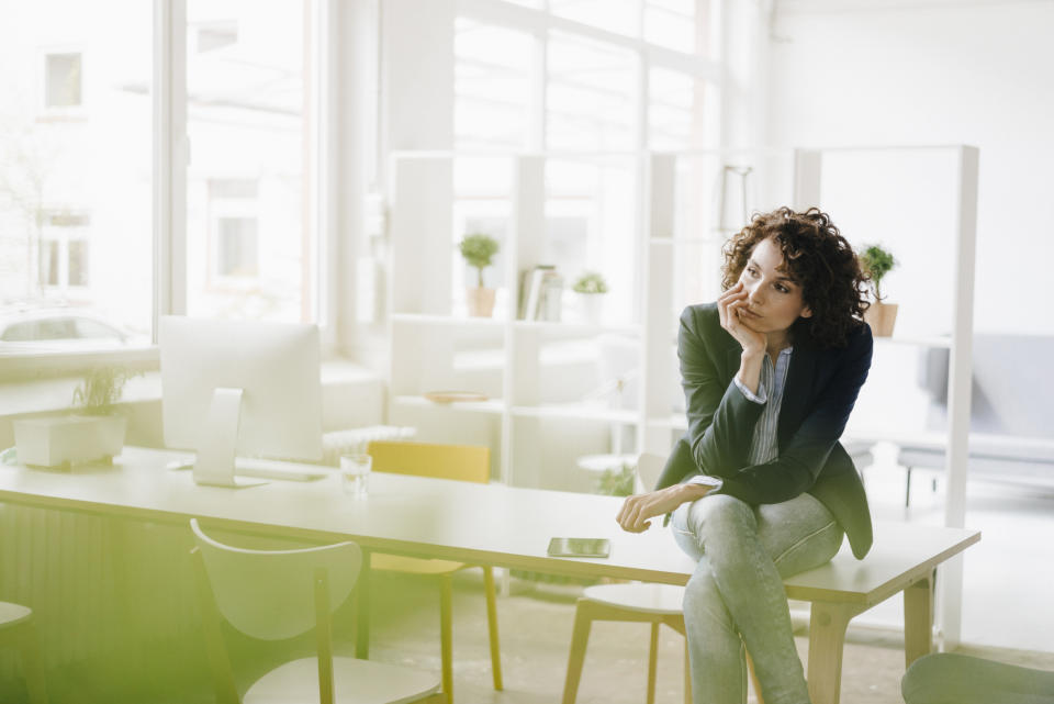 Woman in business attire sitting at office desk, looking contemplative, with a smartphone on the table