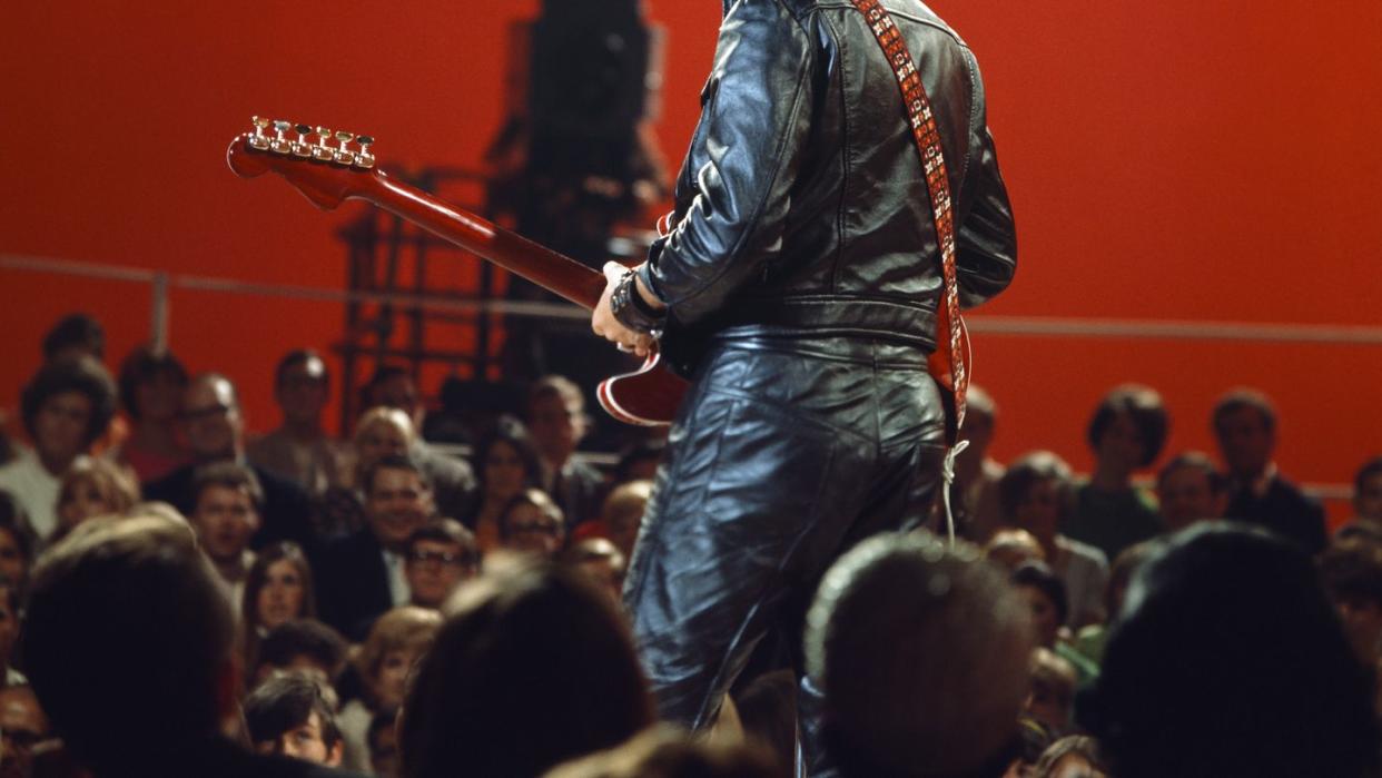 elvis presley smiles as he stands on an elevated stage and a crowd watches him, he wears a black leather jacket and black leather pants and holds his guitar with a strap around his back