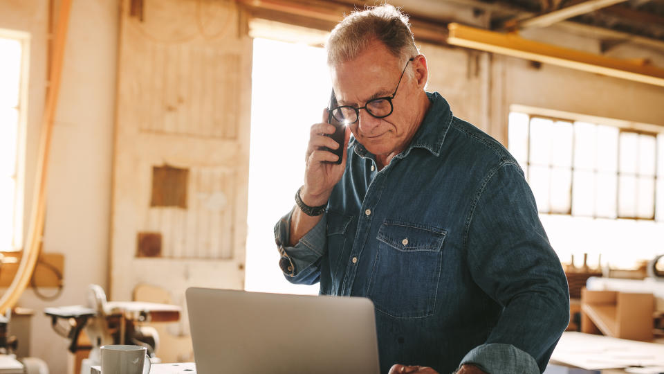 Senior man talking on cell phone and using laptop on work table.