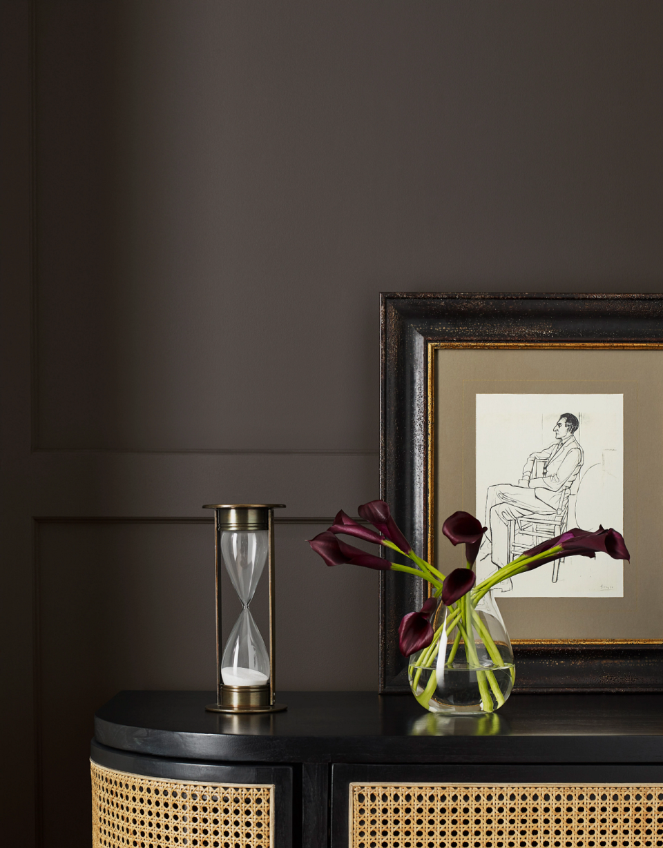 A room painted with dark brown-black walls with wall panelling and a credenza decorated with flowers in a vase