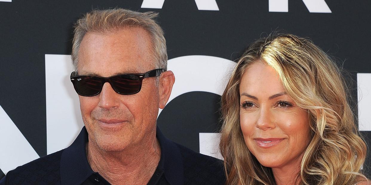 Kevin Costner wearing a black shirt with blacked-out sunglasses while standing next to Christine Baumgartner