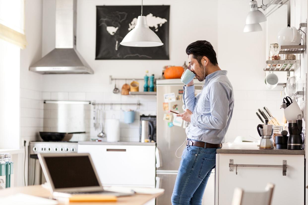 man drinking coffee and listening to phone with headphones in kitchen while taking a break from laptop on table