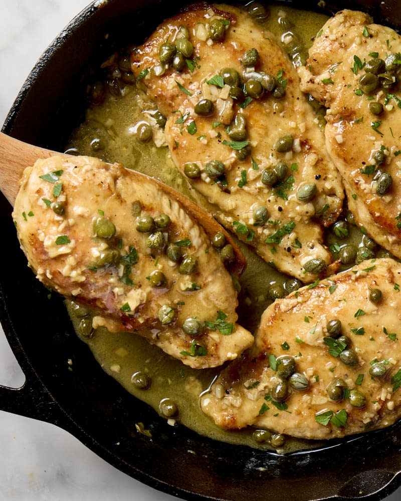 overhead shot of chicken piccata in a cast iron pan, with a piece of chicken being pulled from the pan with a wooden spoon.