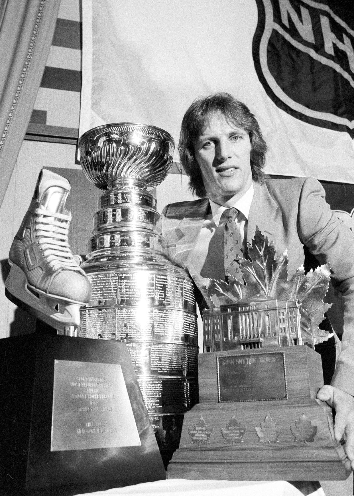 FILE - New York Islanders right wing Mike Bossy poses in New York with some of the spoils reaped after the Islanders defeated the Vancouver Canucks to win the Stanley Cup at the Nassau Coliseum in Uniondale, N.Y., May 20, 1982. Bossy scored two goals in the game and finished the playoffs with 17, earning the Conn Smythe MVP trophy at right and the Sport Magazine MVP trophy, left, At center is the Stanley Cup. Bossy, one of hockey’s most prolific goal-scorers and a star for the New York Islanders during their 1980s dynasty, died Thursday, April 14, 2022, after a battle with lung cancer. He was 65. (AP Photo/Marty Lederhandler, File)