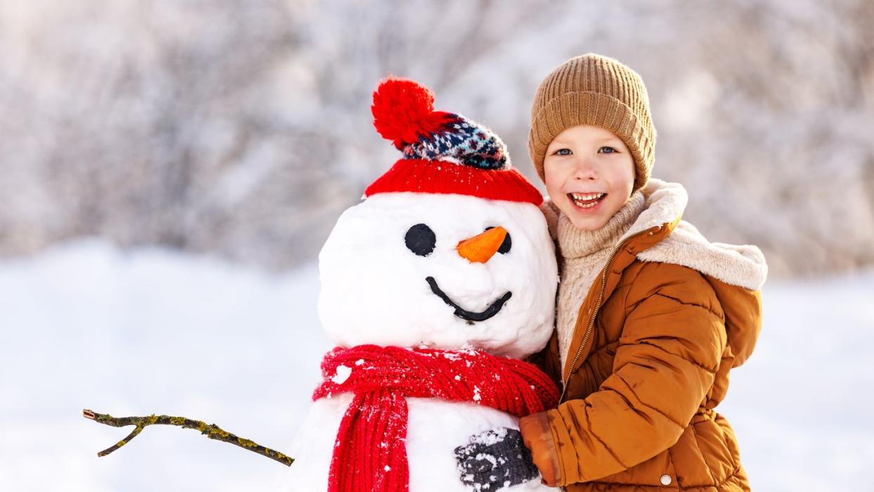 happy child cute little boy in warm clothes with snowman in snowy winter park on beautiful sunny frosty day, selective focus snow games for children, outdoor wintertime activities for kids