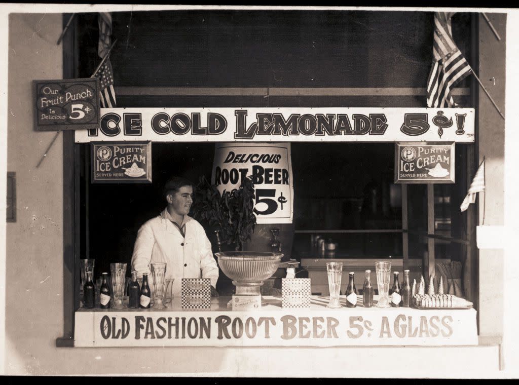 An unidentified man stands behind the counter of a stall that offers lemonade, root beer, Cracker Jacks, and ice cream for sale, 1920s or 1930s.