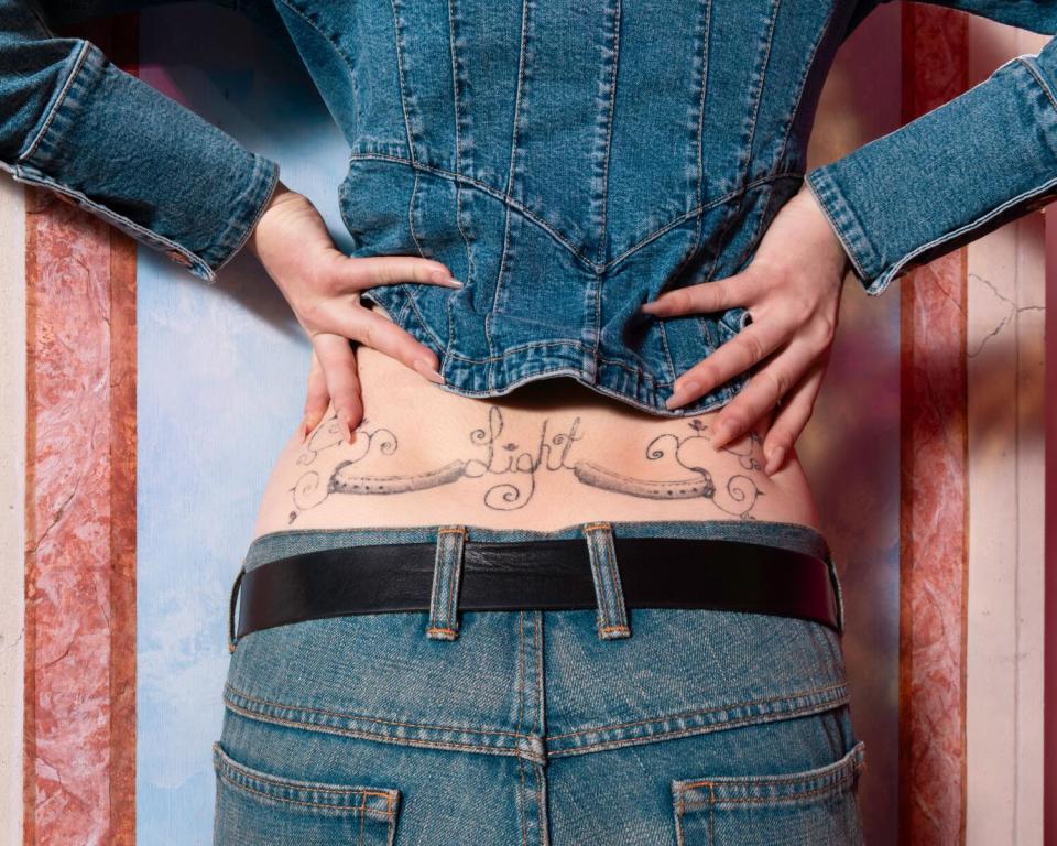 A close-up photo of a model lifting her jean jacket and revealing a tattoo that says "Light" on her back.