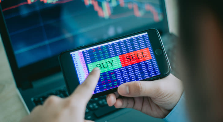 Stocks to buy: smartphone with the words "buy" and "sell" displayed on the screen. The user's finger is about to press buy. Stock charts are in the background of the image.